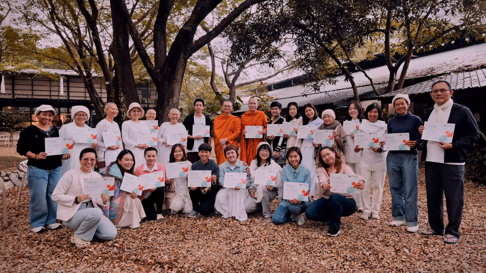 Group of people outdoors holding certificates, with two monks in orange robes at the center, under large trees.