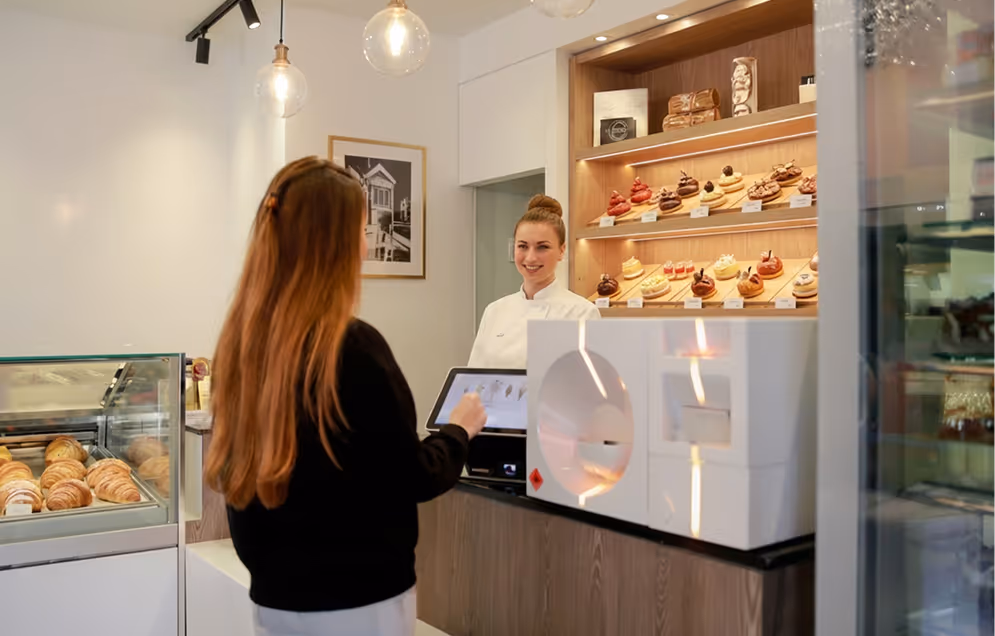 Customer using touchscreen to order at a bakery counter with pastries displayed behind the cashier.