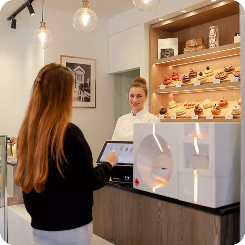 Customers in line at a modern bakery counter with a smiling male baker behind a glass display of croissants and pastries.