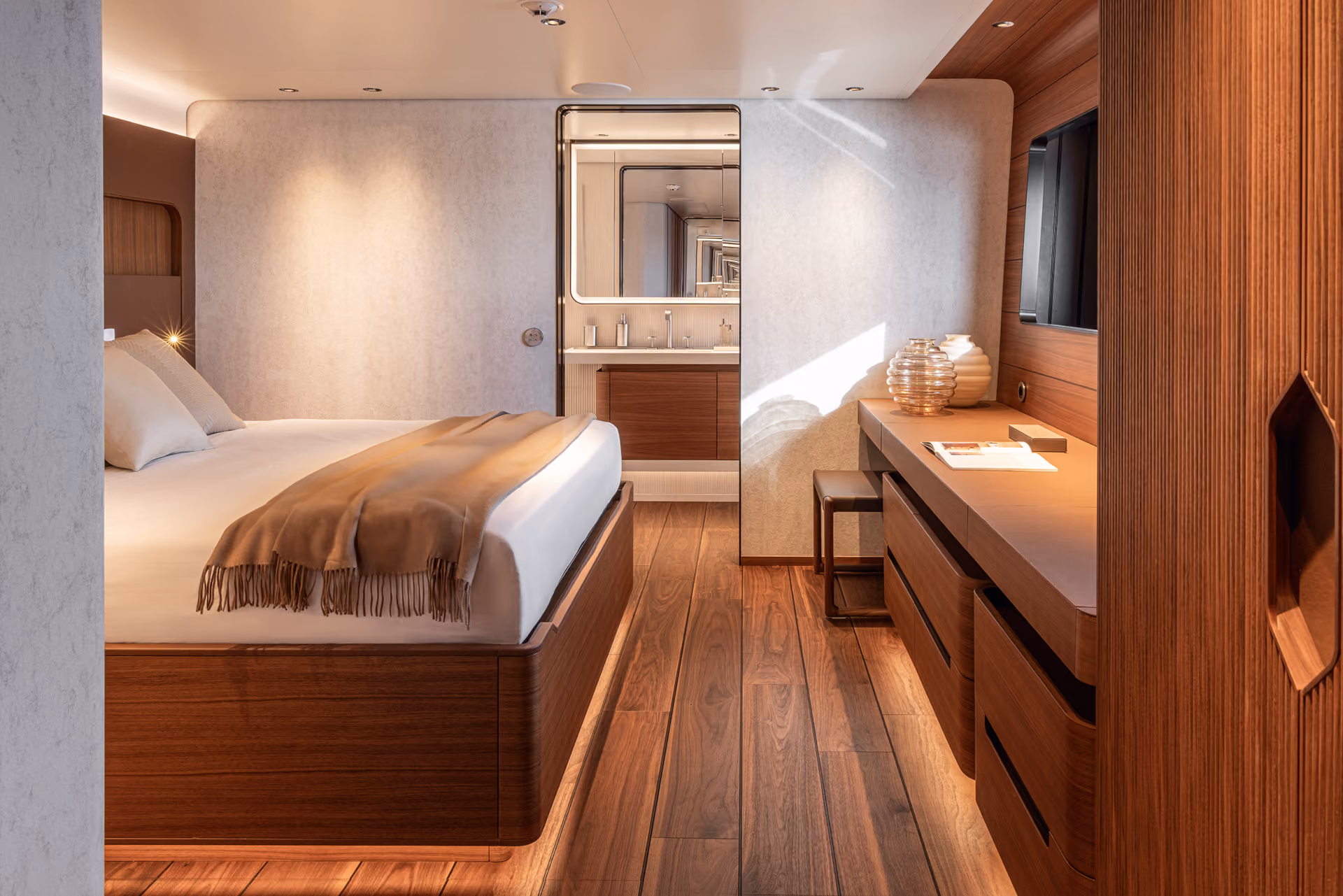 Modernes Schlafzimmer einer Yacht mit Holzfußboden, großem Bett mit weißer Bettwäsche und brauner Decke, Schreibtisch mit Dekoration und Fernseher an der Wand.