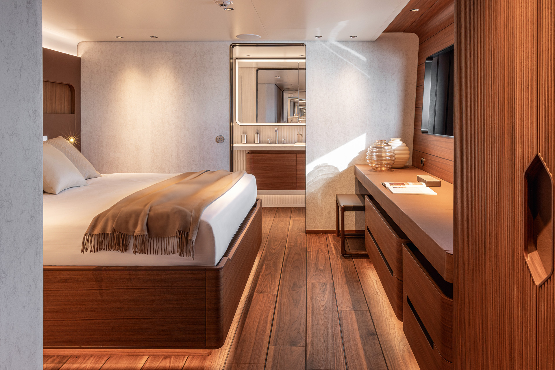 Modernes Schlafzimmer einer Yacht mit Holzfußboden, großem Bett mit weißer Bettwäsche und brauner Decke, Schreibtisch mit Dekoration und Fernseher an der Wand.