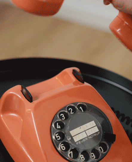 Nahaufnahme eines orangefarbenen Wählscheibentelefons mit einer Hand, die den Hörer anhebt.