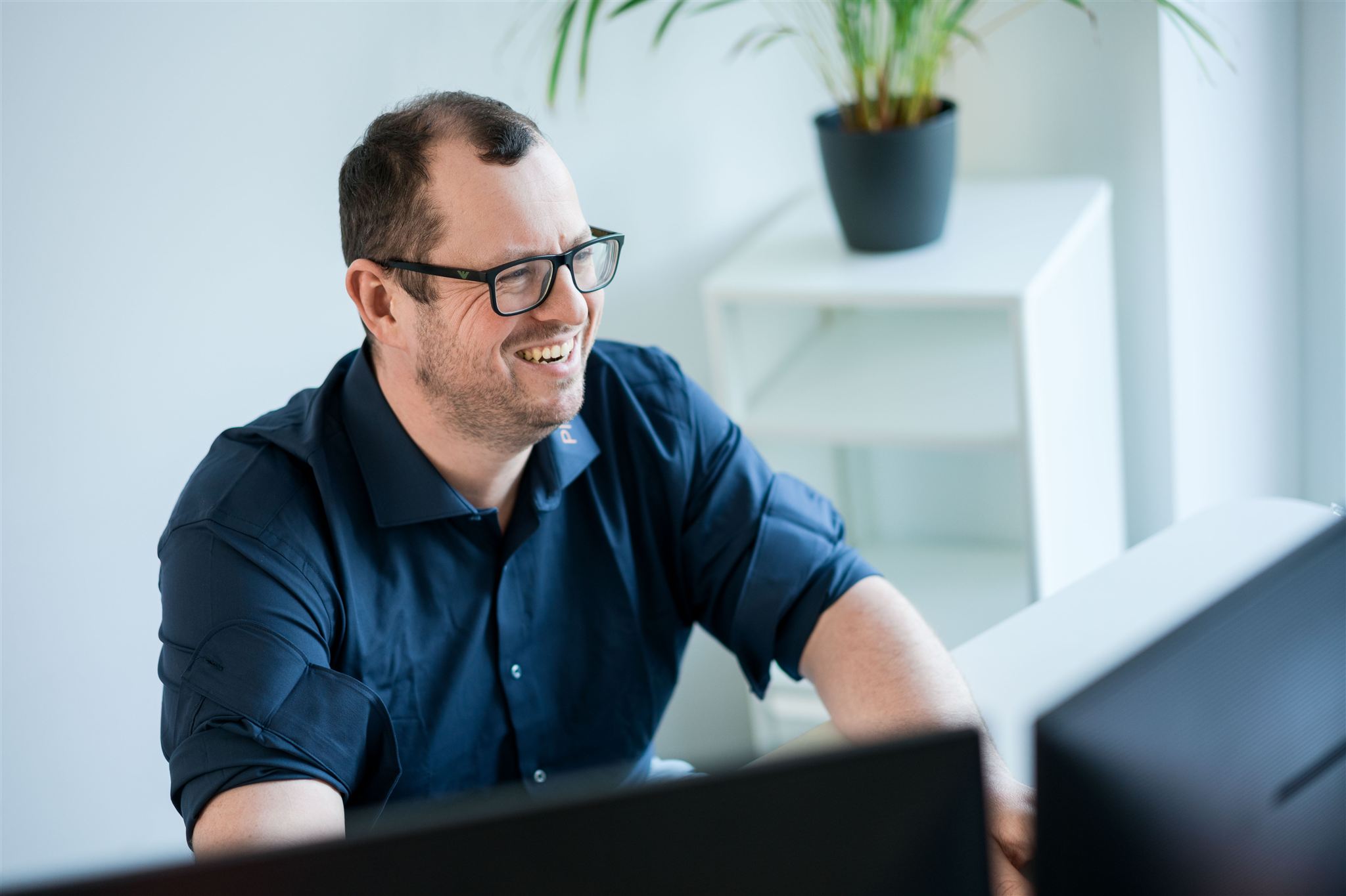 Lächelnder Mann mit Brille und dunklem Hemd, der am Schreibtisch vor Monitoren sitzt.