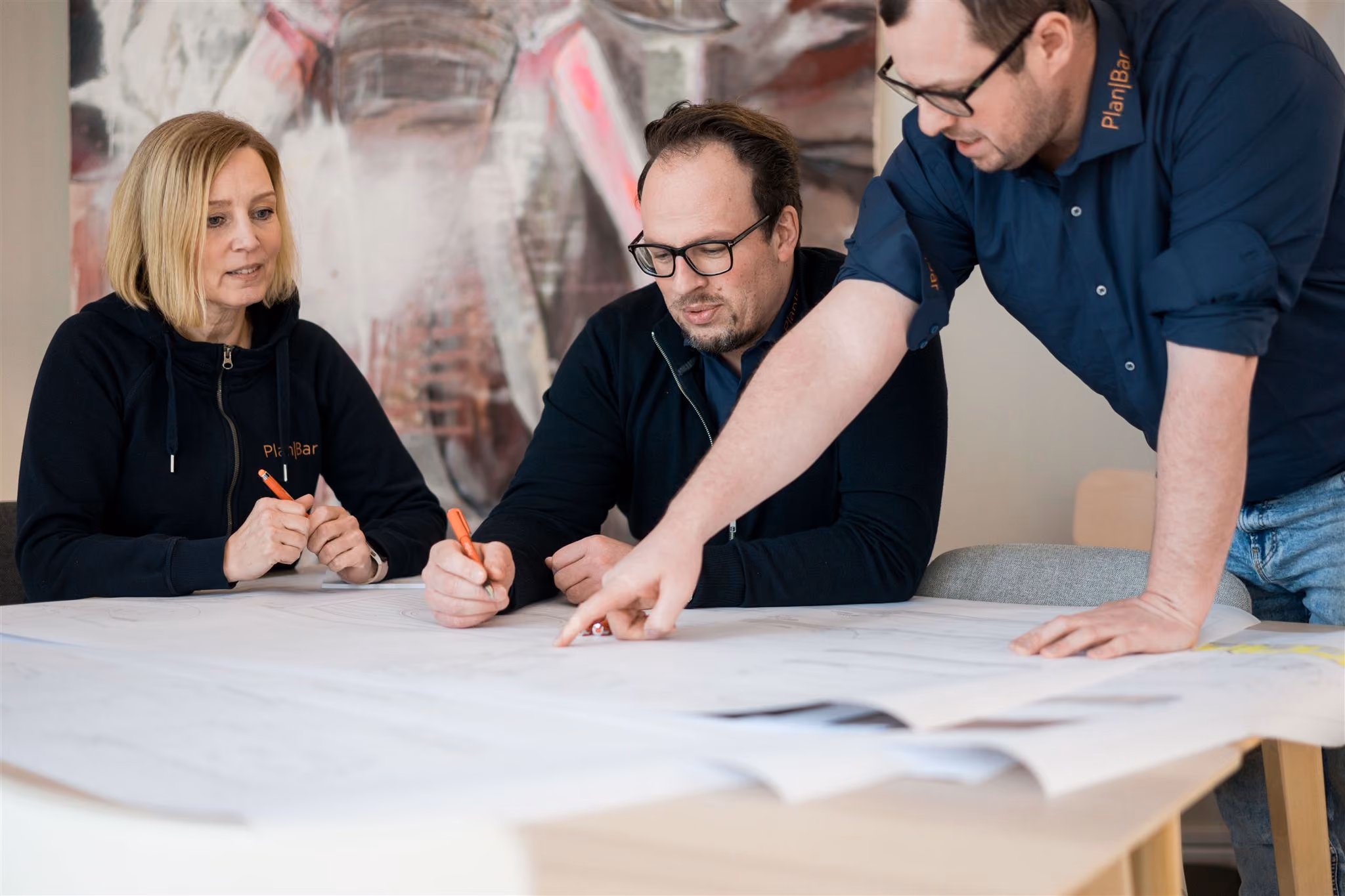 Drei Personen in PlanBar Kleidung besprechen und zeichnen auf großen Architekturplänen an einem hellen Tisch.
