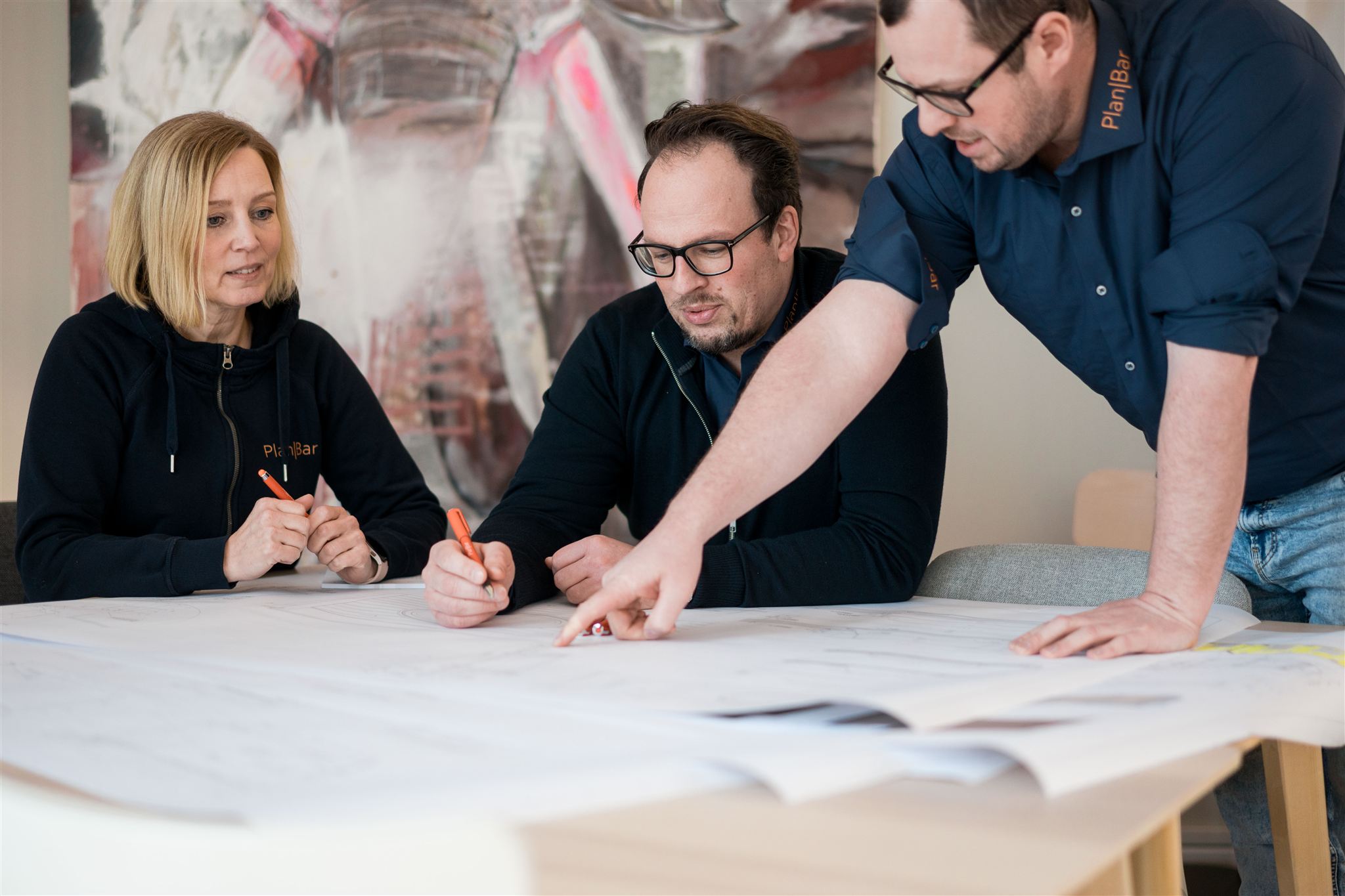 Drei Personen in PlanBar Kleidung besprechen und zeichnen auf großen Architekturplänen an einem hellen Tisch.