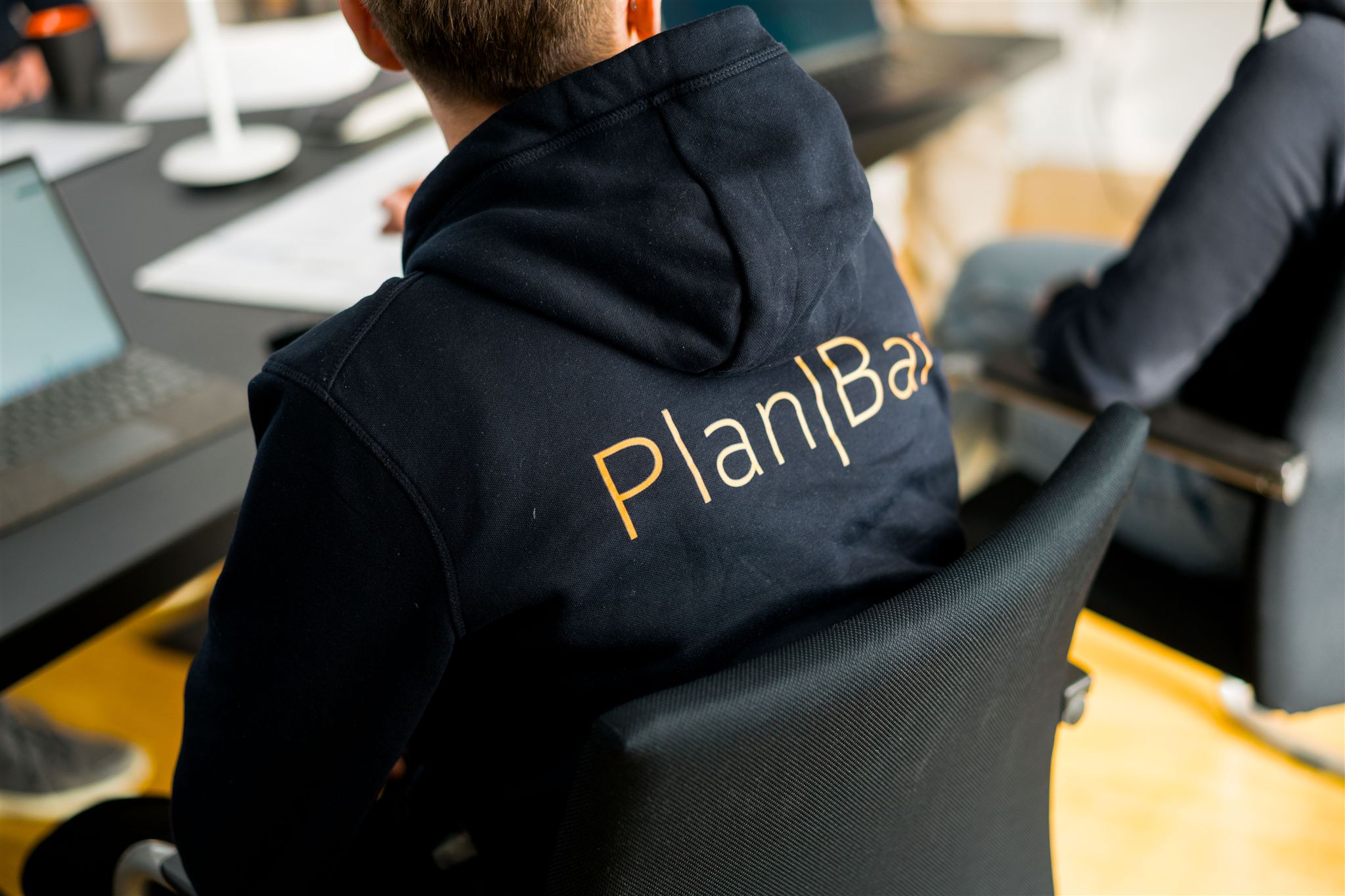 Person wearing a navy blue hoodie with the word PlanBar printed on the back, sitting at a desk with a laptop.