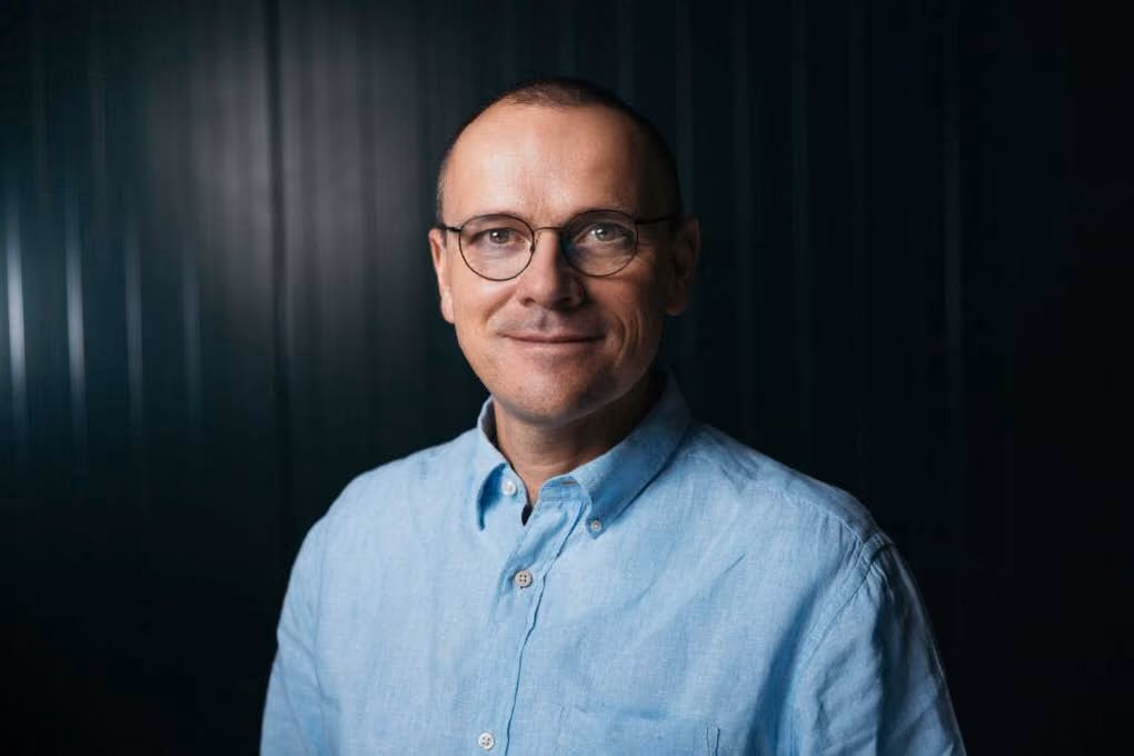 Mann mit Brille und blauem Hemd vor dunklem Hintergrund lächelt in die Kamera.