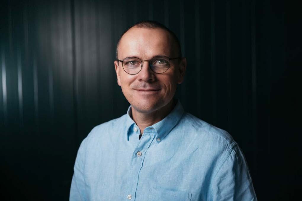 Mann mit Brille und blauem Hemd vor dunklem Hintergrund lächelt in die Kamera.