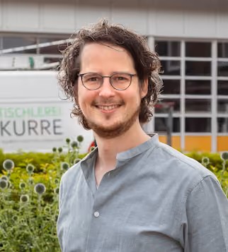 Porträt eines lächelnden Mannes mit Brille, lockigem Haar und grauem Hemd vor einer Werkstatt.
