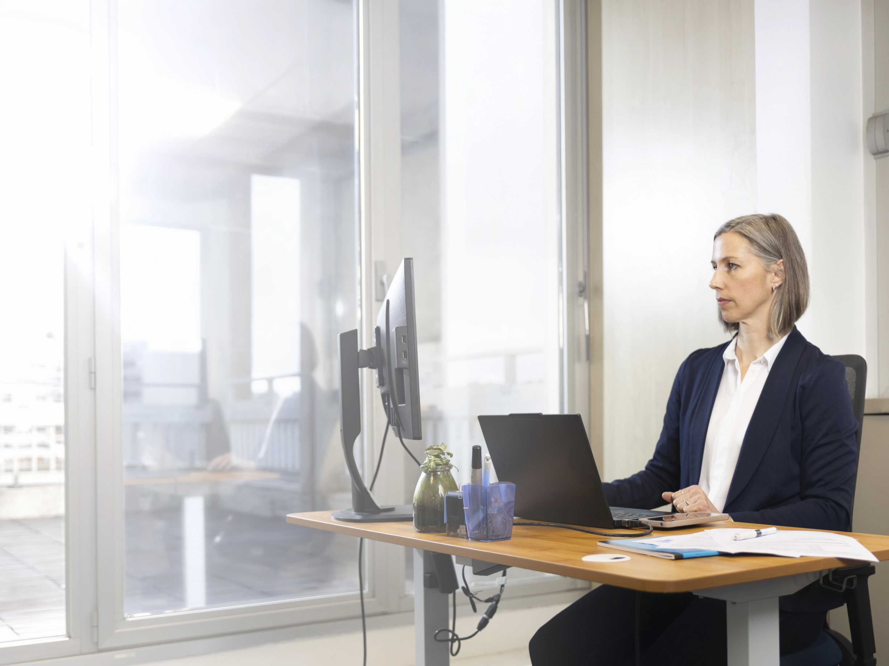 Femme professionnelle assise à un bureau, travaillant sur un ordinateur portable avec un écran externe dans un bureau lumineux.