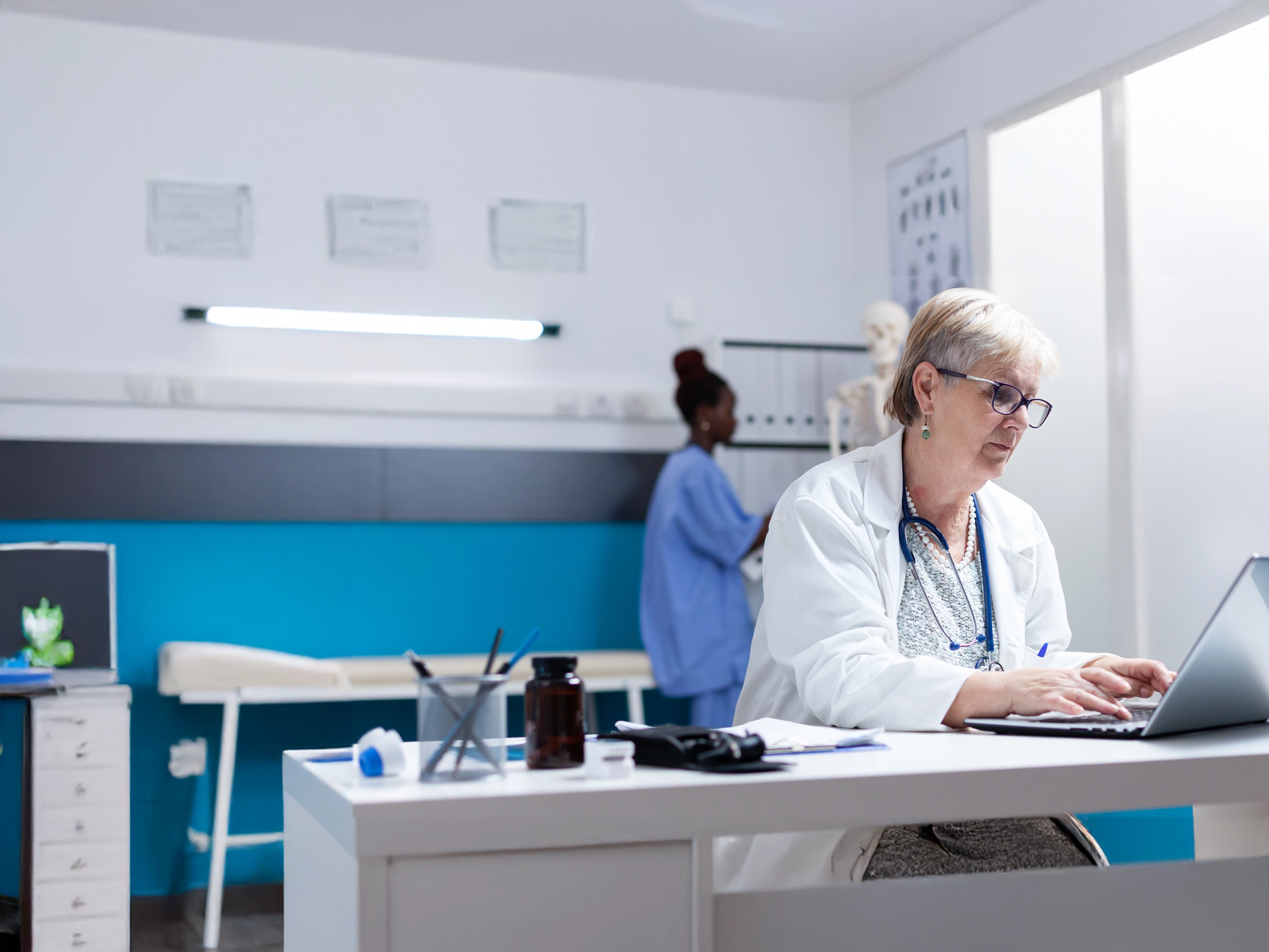 Médecin âgée tapant sur un ordinateur portable dans un cabinet médical avec une infirmière debout en arrière-plan.
