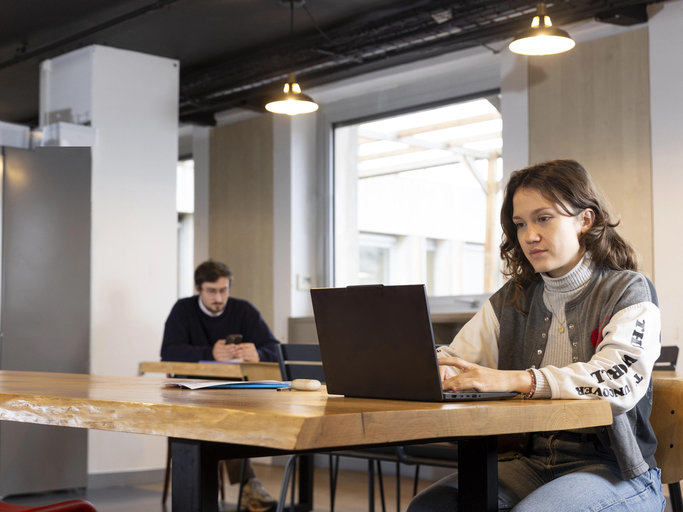 Jeune femme travaillant sur un ordinateur portable à une grande table en bois dans un espace de travail partagé avec un homme en arrière-plan utilisant son téléphone.