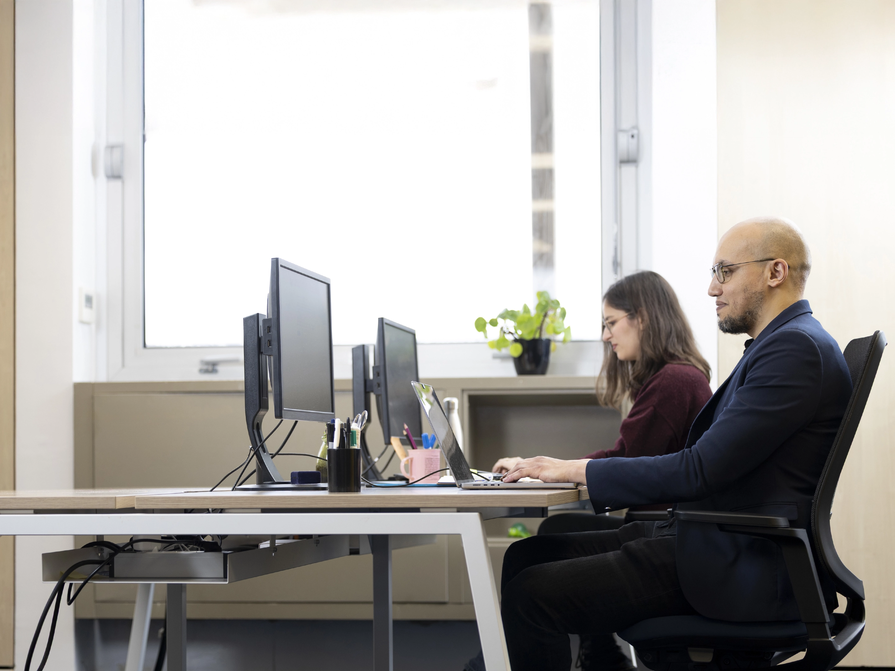 Deux professionnels travaillant côte à côte sur des ordinateurs dans un bureau lumineux avec une grande fenêtre.