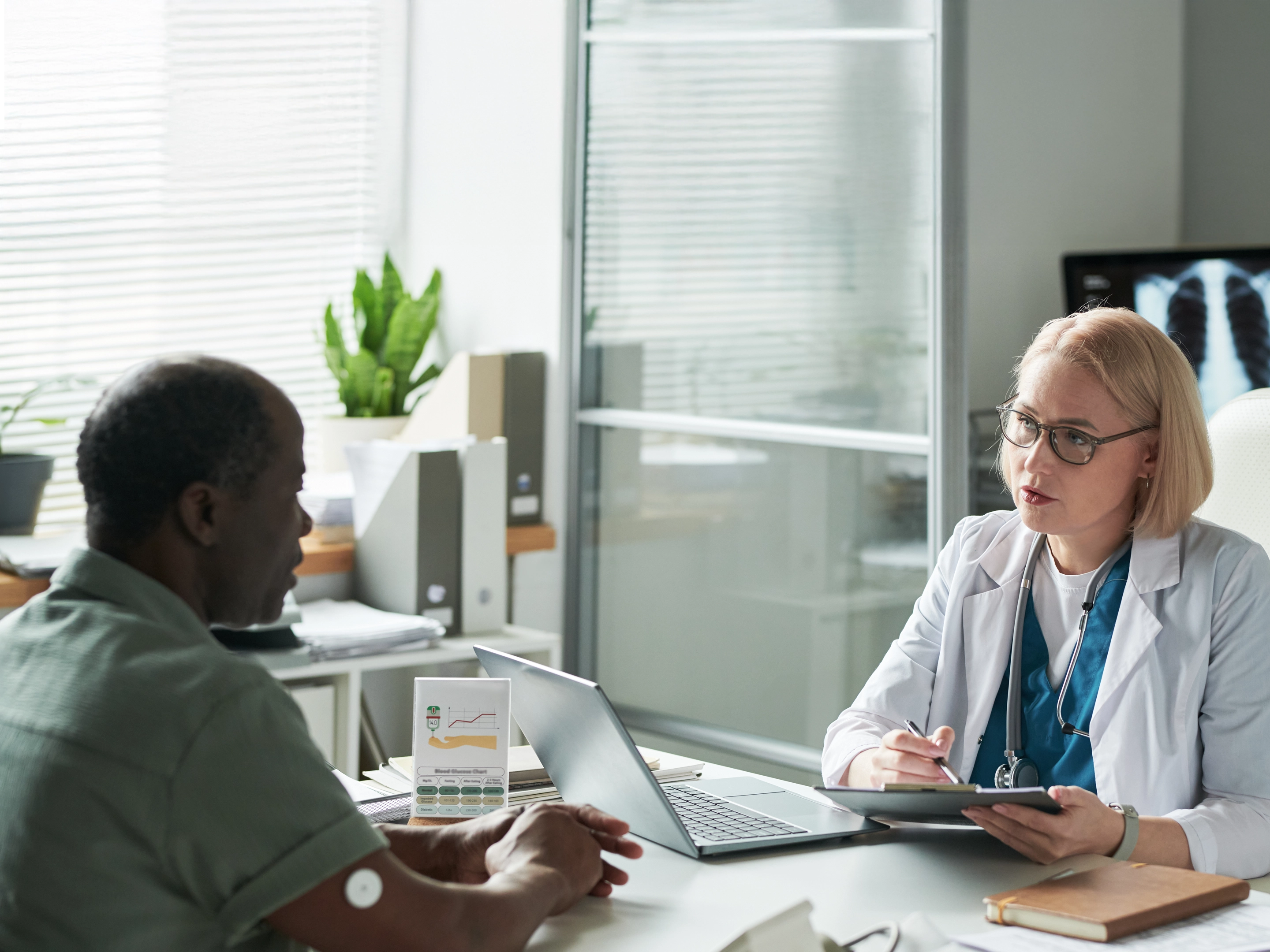Une médecin consulte un patient assis de l'autre côté d'un bureau dans un cabinet médical lumineux.