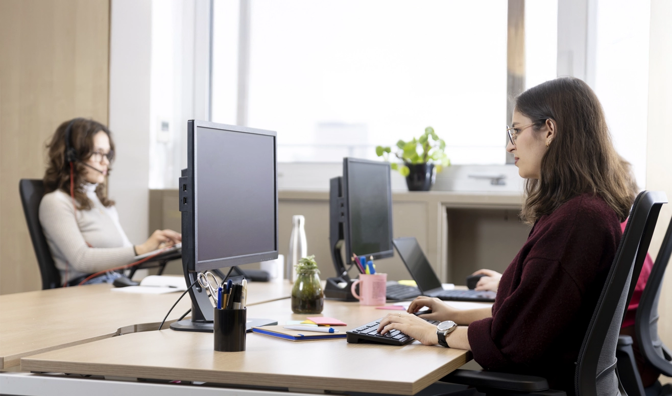 Deux femmes travaillant sur ordinateur dans un bureau moderne avec des plantes et des fournitures de bureau sur les bureaux.