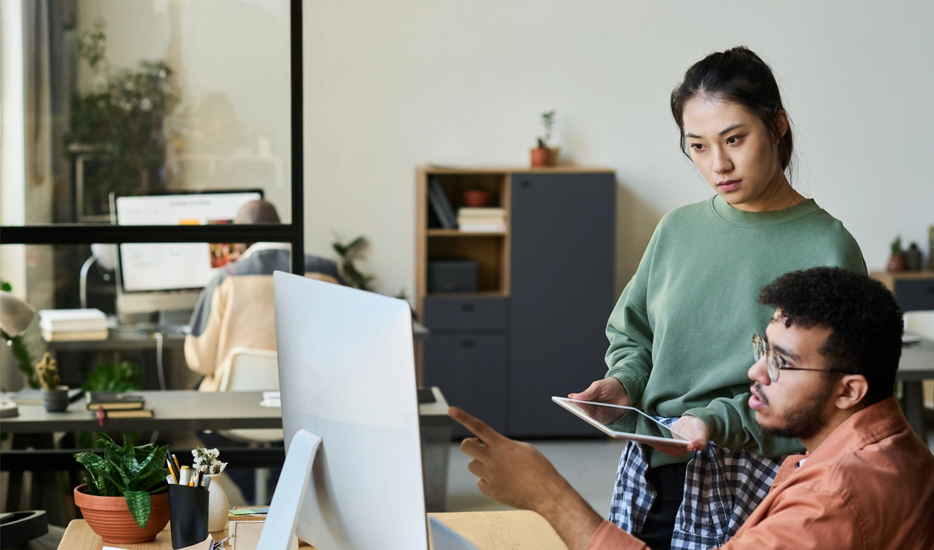 Deux collègues discutent devant un ordinateur dans un bureau moderne, une femme tenant une tablette et un homme pointant l'écran.