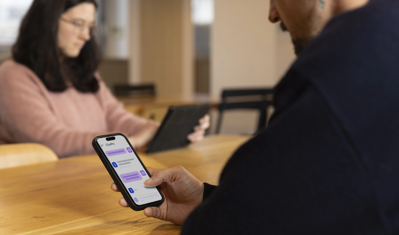 Un homme utilise un smartphone affichant une conversation de chat tandis qu'une femme à l'arrière-plan consulte une tablette.