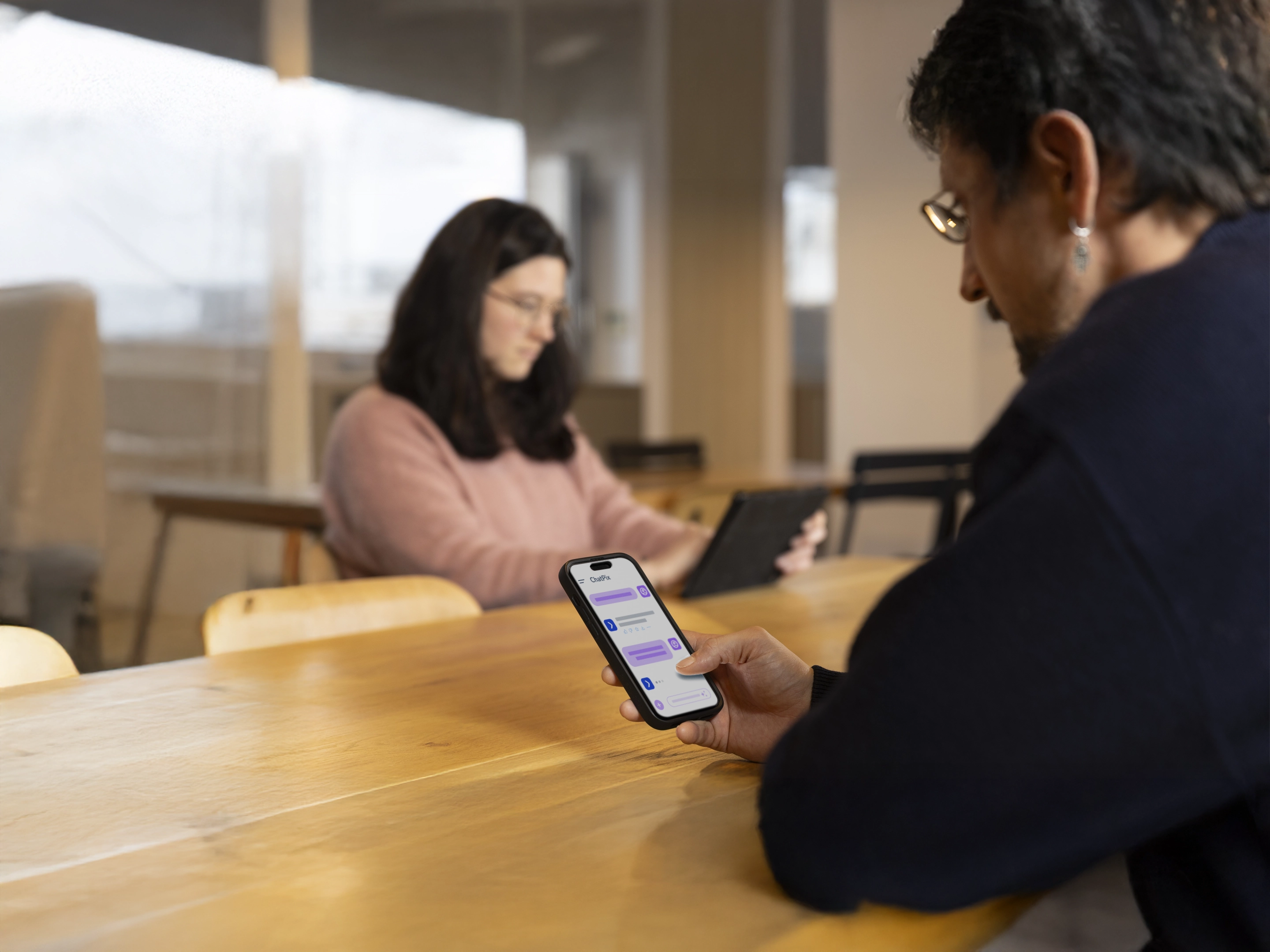 Un homme utilise un smartphone affichant une conversation avec ChatGPT tandis qu'une femme consulte une tablette à l'arrière-plan dans une salle lumineuse.