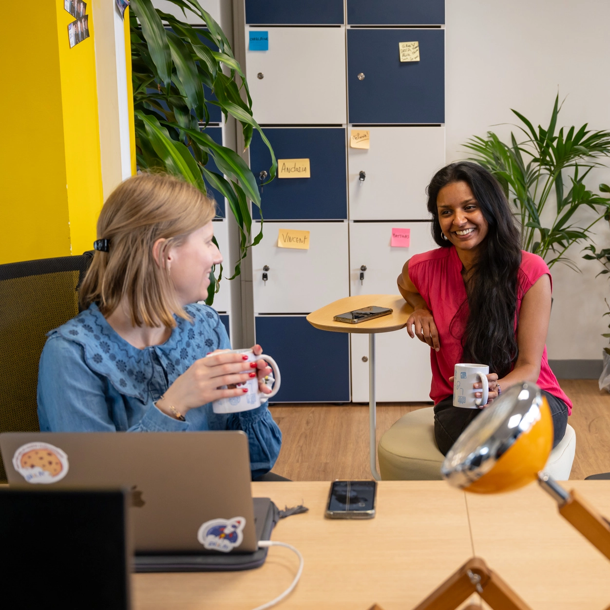 Deux femmes souriantes discutent en tenant des tasses de café dans un bureau avec des casiers bleus et blancs en arrière-plan dans les bureaux de Pix 