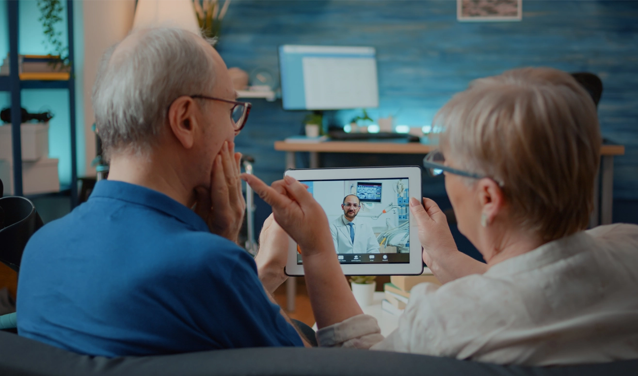 Un couple âgé consulte un médecin via une téléconsultation sur une tablette dans un salon moderne.