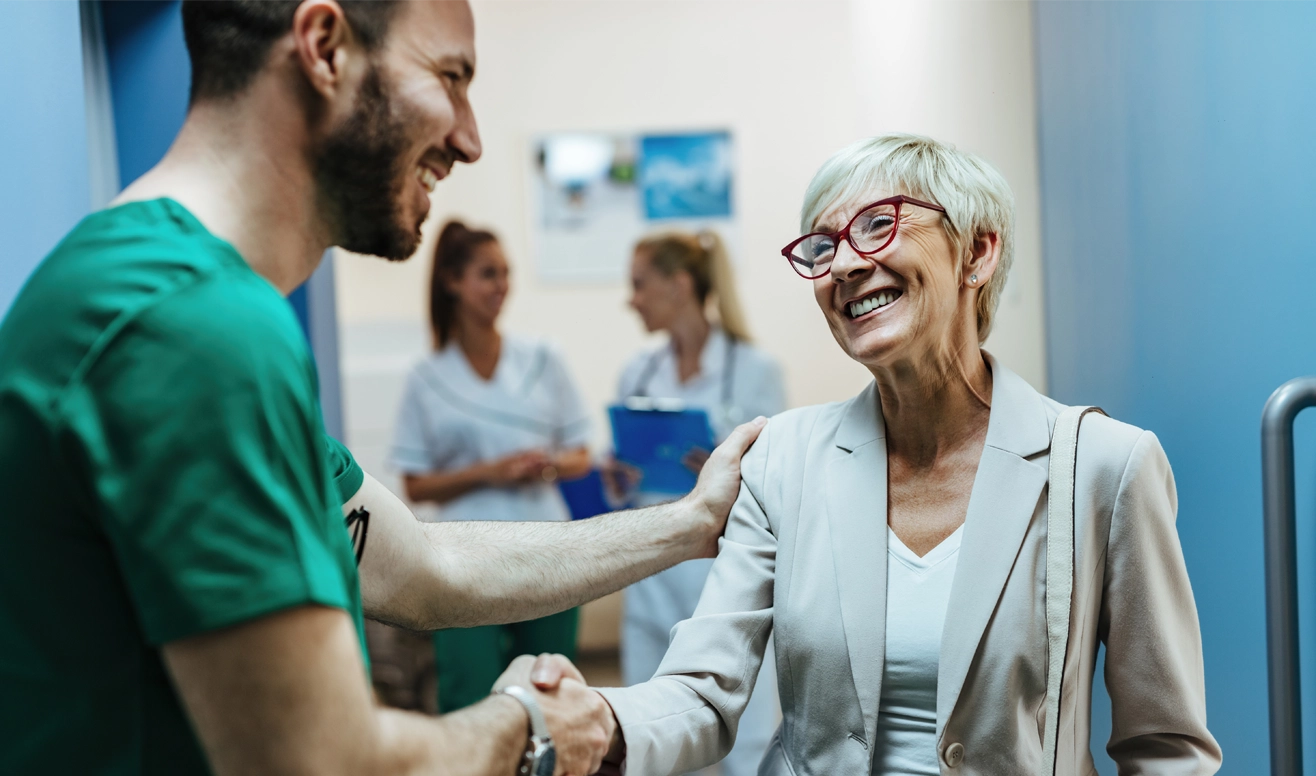 Un professionnel de santé serrant la main et réconfortant une femme âgée souriante dans un couloir d'hôpital.