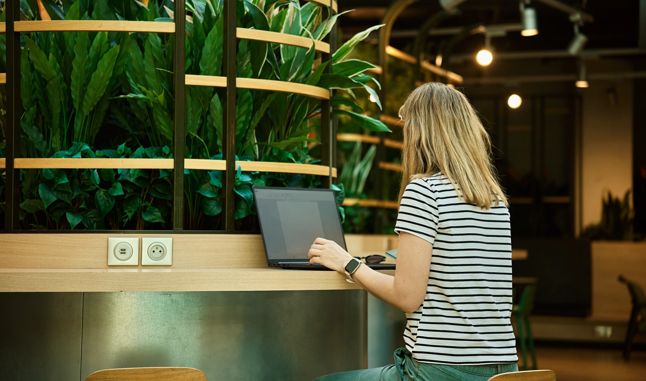 Femme assise à un bureau avec un ordinateur portable, entourée de plantes vertes dans un espace intérieur lumineux.