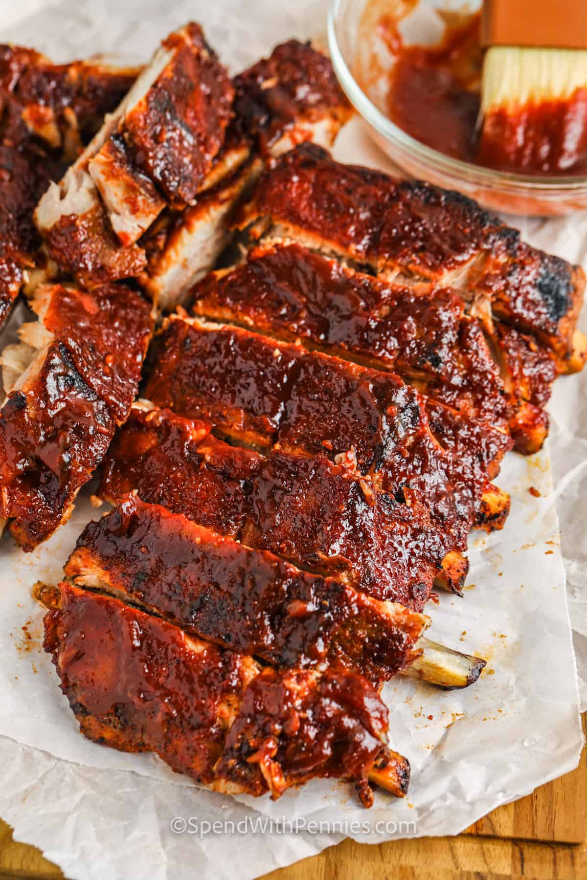 Barbecue pork ribs sliced and glazed with rich barbecue sauce on parchment paper with a brush and sauce bowl nearby.