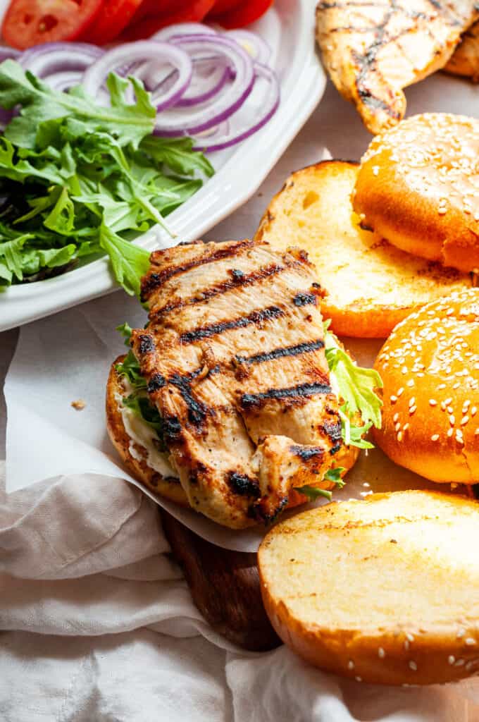 Grilled chicken sandwich with lettuce on a sesame bun, next to sliced tomatoes, red onions, and arugula on a white plate.