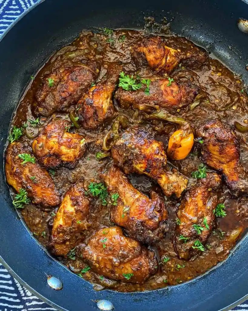 Pan of cooked chicken drumsticks and wings in a dark, savory sauce garnished with chopped herbs.