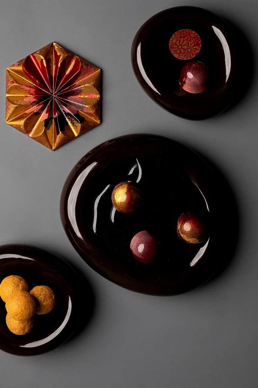 Lśniące ciemne talerze z ozdobnymi kulkami i złoto-czerwonym sześciokątnym dekoracyjnym elementem na szarym tle.