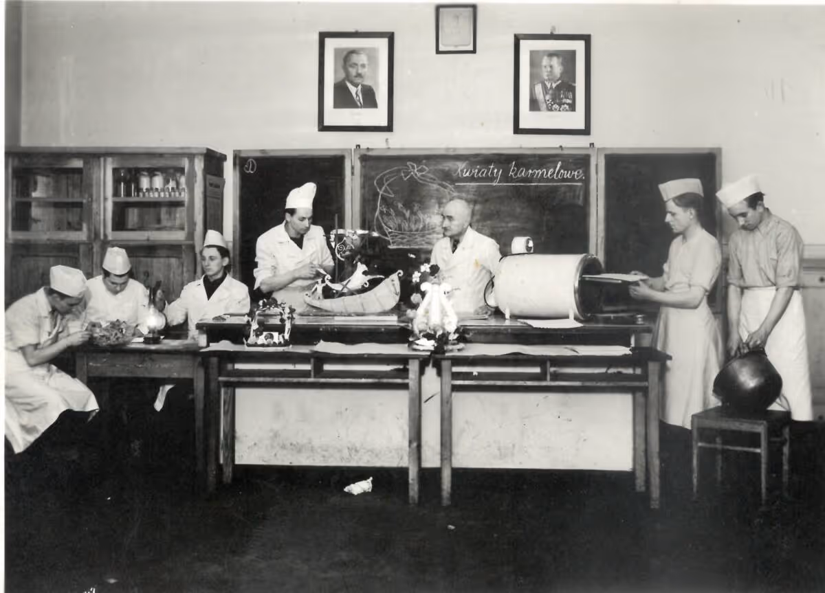 Czarno-białe zdjęcie przedstawiające ośmiu mężczyzn w fartuchach i czapkach kucharskich pracujących przy długim stole w kuchni lub laboratorium cukierniczym.