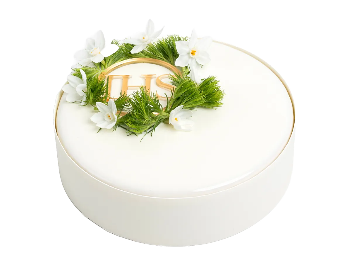 Biały okrągły tort komunijny z dekoracją z zielonych gałązek i białych kwiatów oraz złotym napisem IHS.