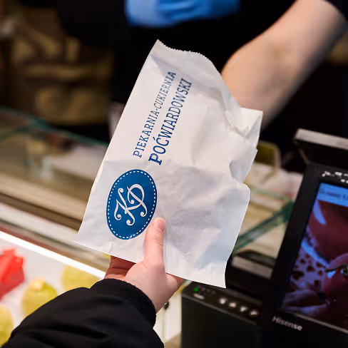 Ręka przekazująca papierową torbę z logo i nazwą piekarnio-cukierni Poćwiardowski przy kasie sklepowej.