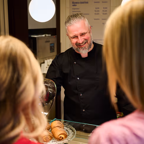 Uśmiechnięty mężczyzna w czarnym fartuchu serwujący rogaliki dwóm kobietom przy ladzie cukierni.