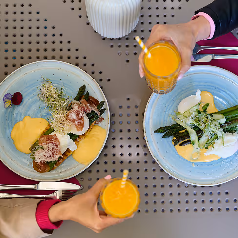 Para trzymająca szklanki z pomarańczowym sokiem nad talerzami z jedzeniem na metalowym stole z otworami.