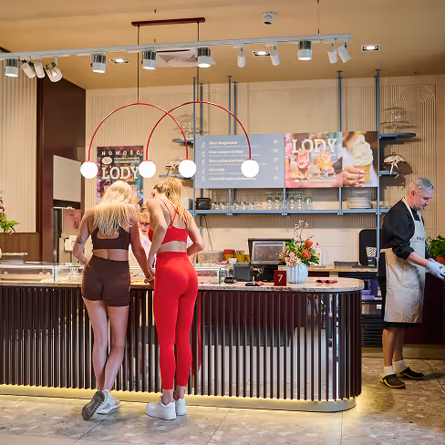 Dwie kobiety w sportowych strojach stoją przy ladzie lodziarni w Piekarnia Cukiernia Powiardowski, a mężczyzna w fartuchu przygotowuje lody w tle.