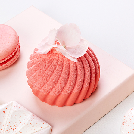 Różowy deser monoporcja z falistą teksturą ozdobiony białym kwiatem na jasnoróżowym tle obok różowego makaronika.