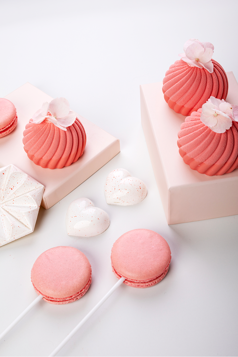 Różowe dekoracyjne słodkości, w tym makaroniki na patyczkach, serduszka i monoporcje ozdobione kwiatami, ułożone na jasnym tle.