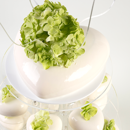Biały tort w kształcie serca z zielonymi kwiatami dekoracyjnymi u góry, umieszczony na szklanym stojaku z dodatkowymi małymi sercowymi monoporcjami i zielonymi kwiatami.