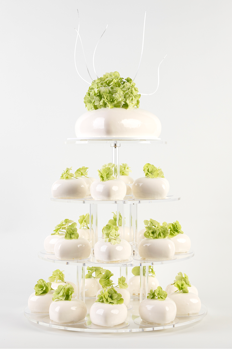 Czteropoziomowy stojak z białymi, błyszczącymi mini tortami ozdobionymi zielonymi kwiatami.