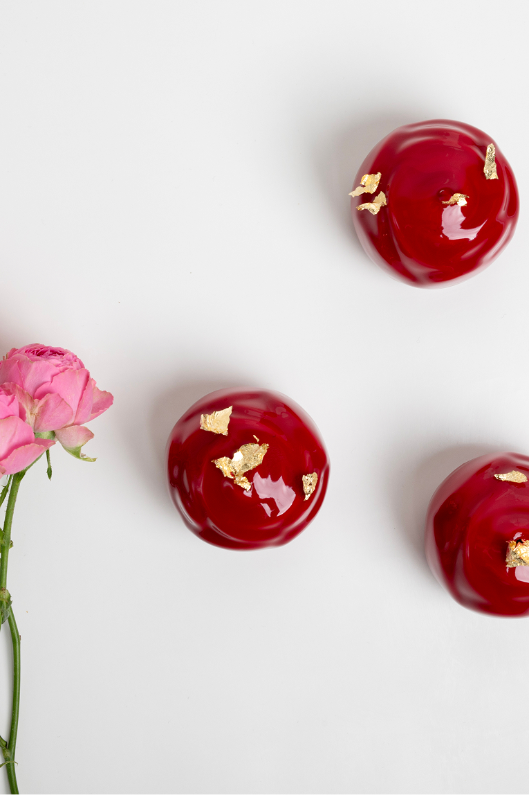 Trzy czerwone desery monoporcje o błyszczącej powierzchni z ozdobami z jadalnego złota obok różowego kwiatu na białym tle.