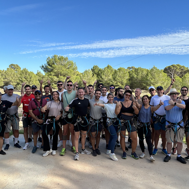 Group of diverse adults wearing safety harnesses posing outdoors under a clear blue sky with trees in the background.