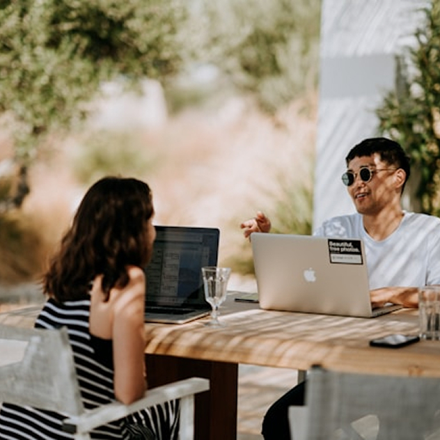 Two people sitting outdoors at a wooden table with laptops, having a conversation in a shaded area.