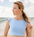 A woman with light brown hair in a ponytail, wearing a light blue sleeveless top, stands on a beach with a gentle ocean breeze. She is looking to the side, with a soft sky and clouds in the background.