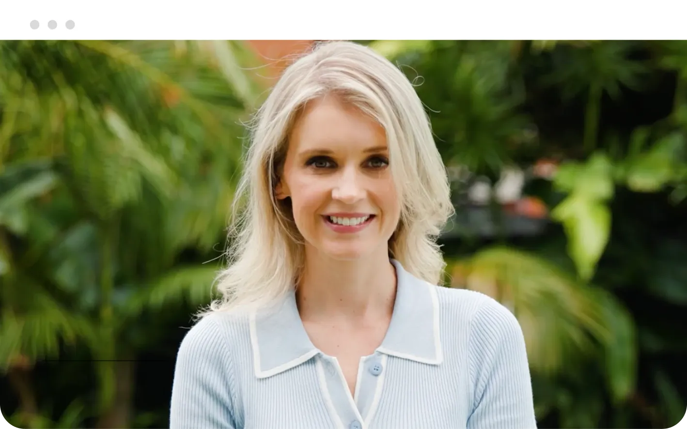 A woman with long, wavy blonde hair, wearing a light blue cardigan, smiling while standing outdoors in a lush garden with green foliage in the background.