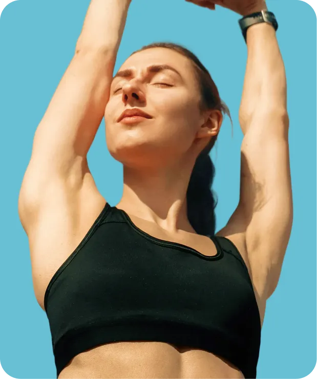 Woman wearing a black sports bra stretching with arms raised, eyes closed, against a blue background.