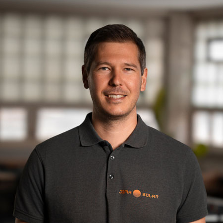 Man wearing a dark polo shirt with JOMA SOLAR logo smiling indoors with blurred windows in the background.
