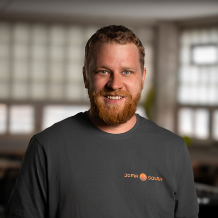 Smiling man with red beard wearing a Joma Solar black T-shirt in an indoor setting.