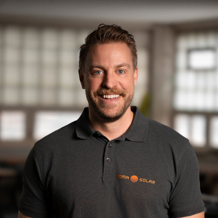 Smiling man with short brown hair and beard wearing a dark polo shirt with Joma Solar logo in an indoor setting.