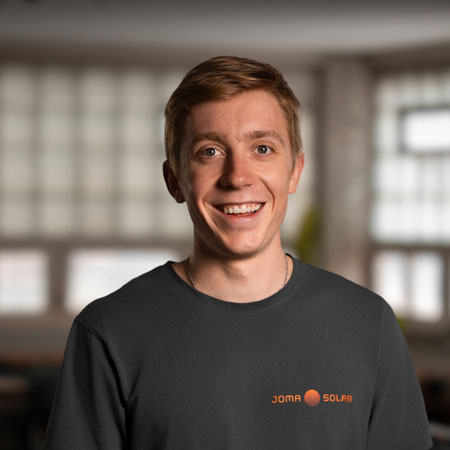Smiling young man with short light brown hair wearing a dark JOMA SOLAR shirt in a softly lit indoor space.